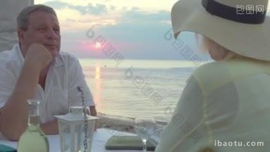 老年夫妇在户外度过浪漫的夜晚，他们坐在日落时分的海滩上的餐厅里，喝着白葡萄酒